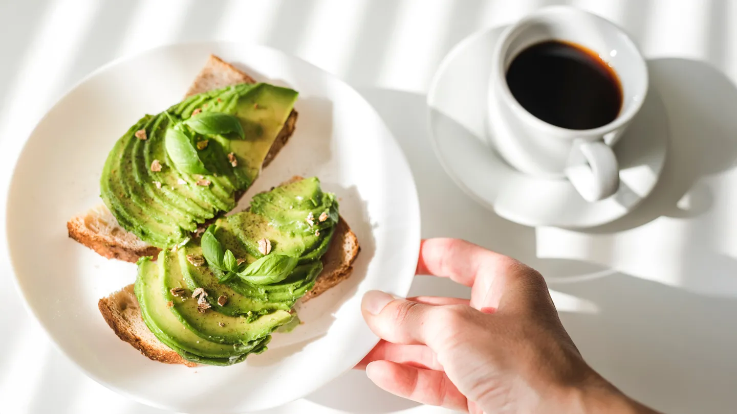 man reaching for a plate of avocado toast next to mug of black coffee