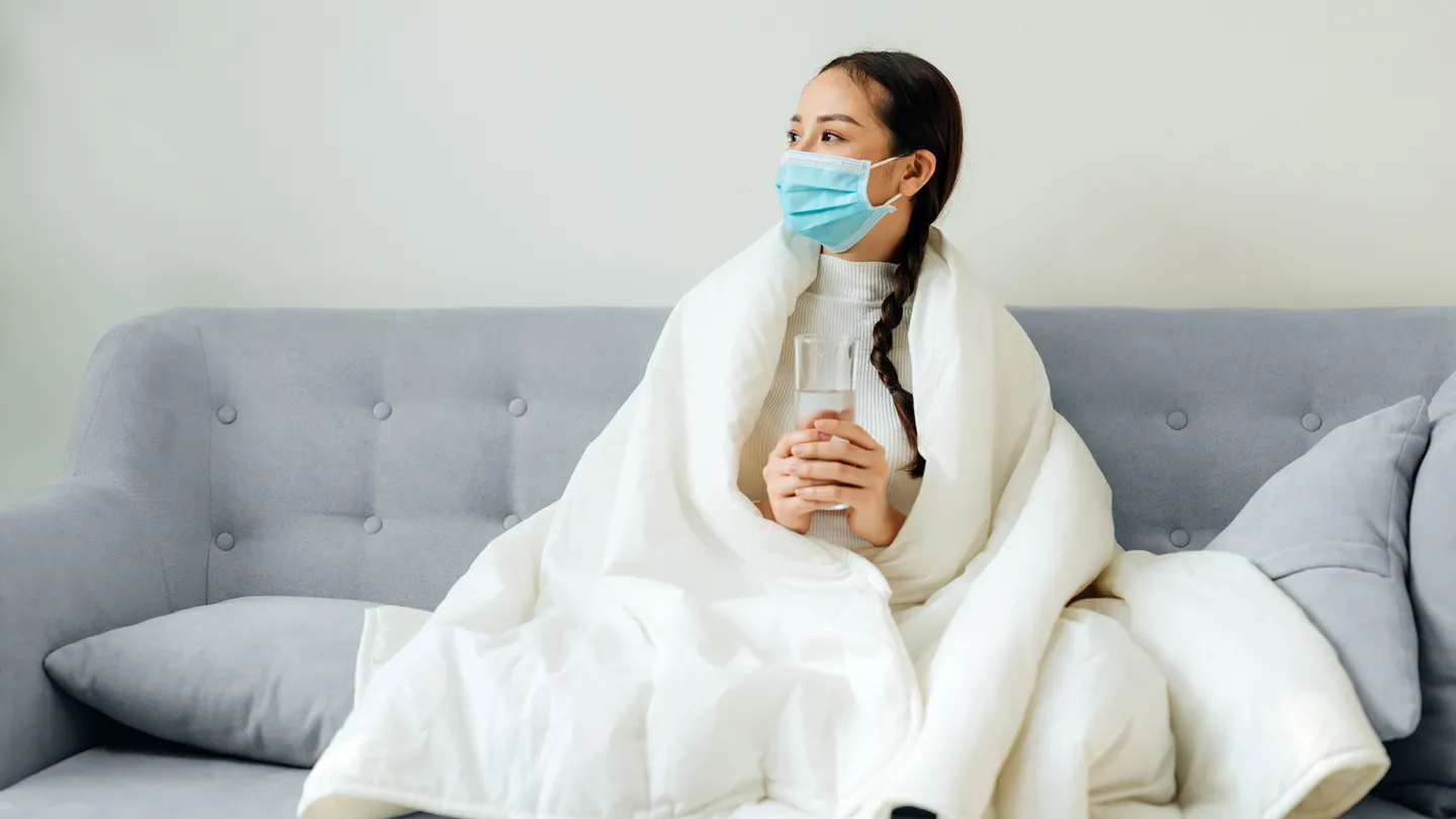 Woman sitting on sofa indoors wearing a mask holding a glass of water