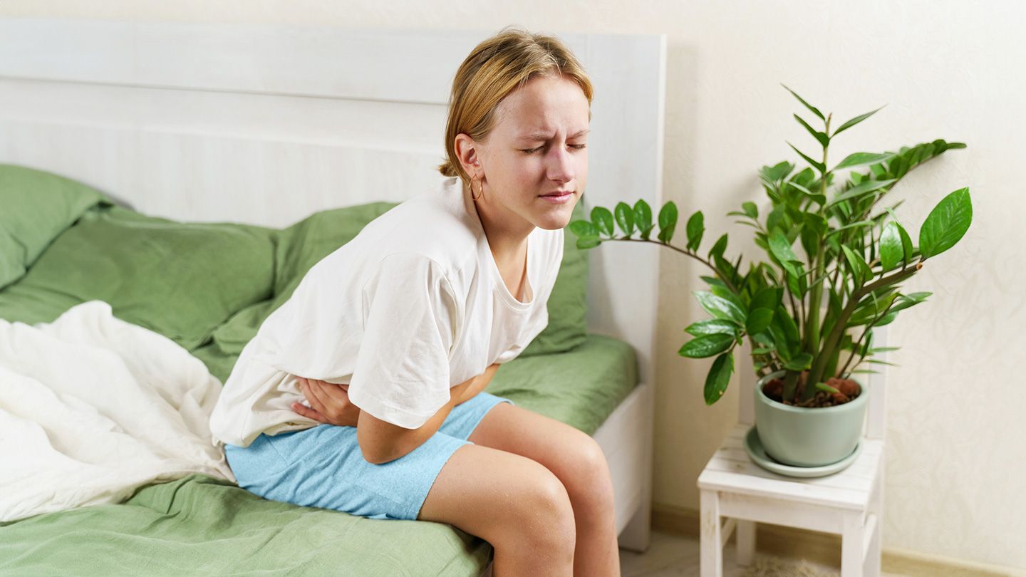 pained face woman with stomach pain sitting on edge of bed green sheets blue shorts plant on chair