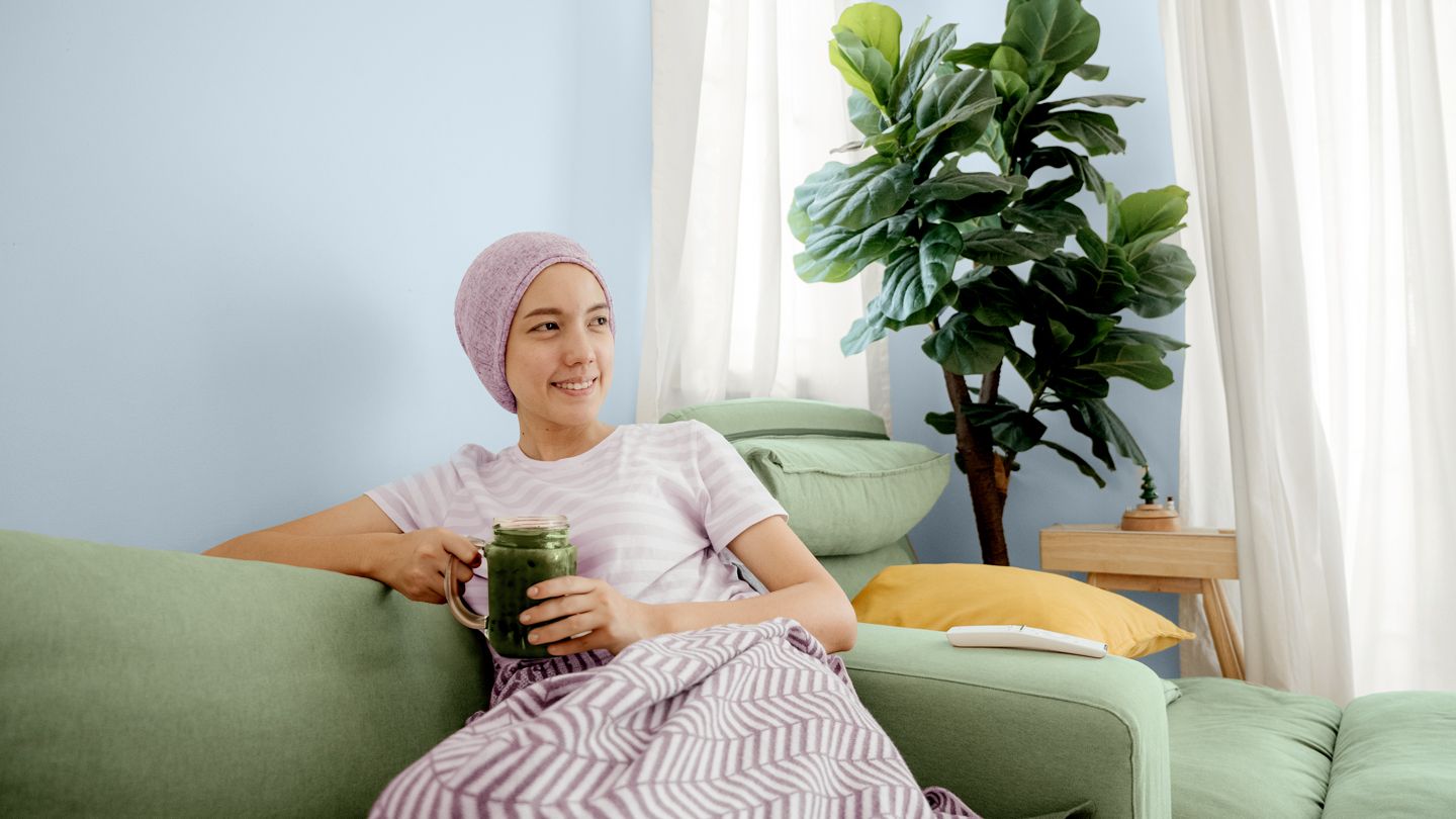 young woman going through chemotherapy sitting on counch drinking a green smoothie