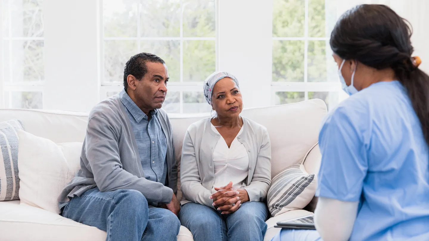 mature Black woman with cancer sitting with husband on couch speaking with healthcare professional