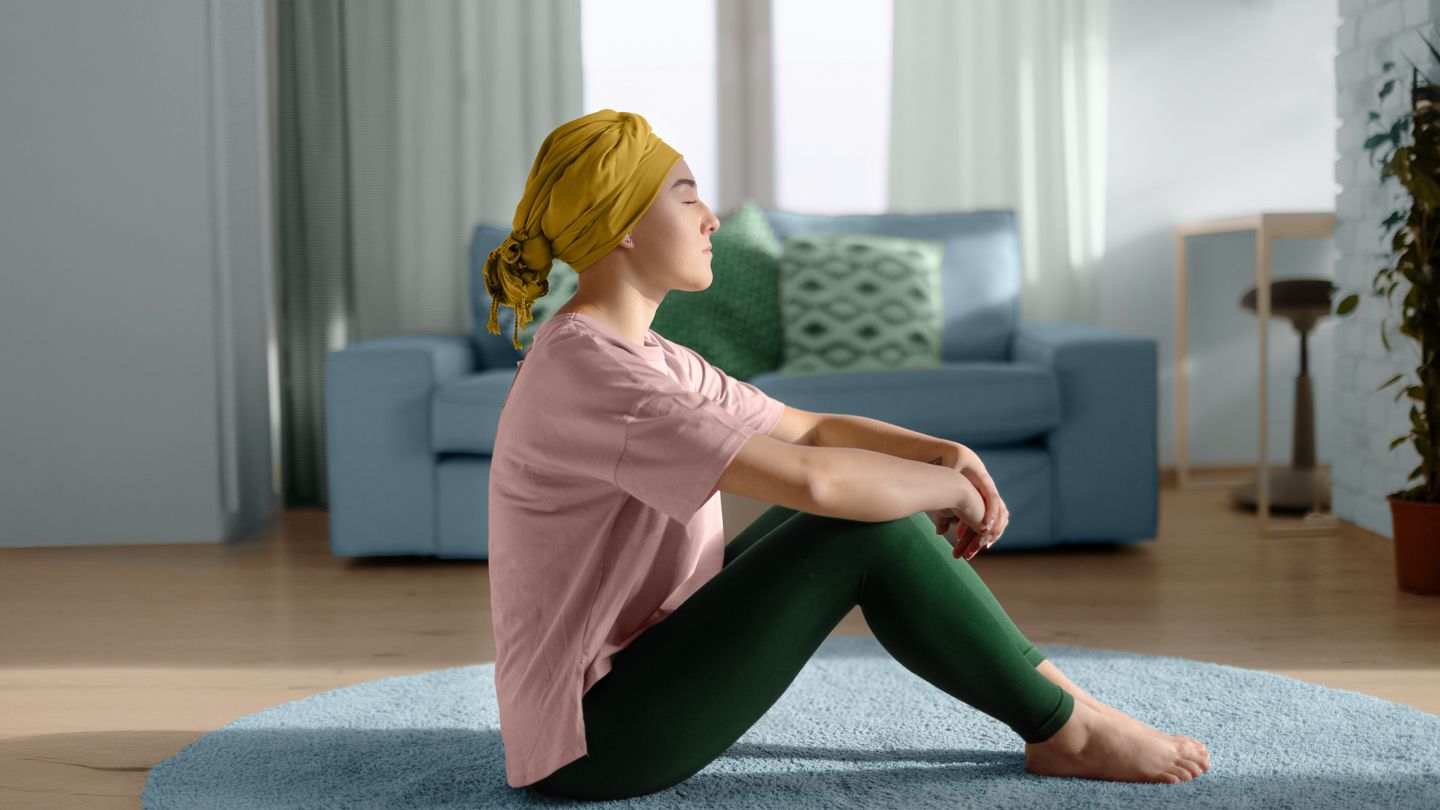 young woman wearing chemotherapy scarf on her head meditating in living room