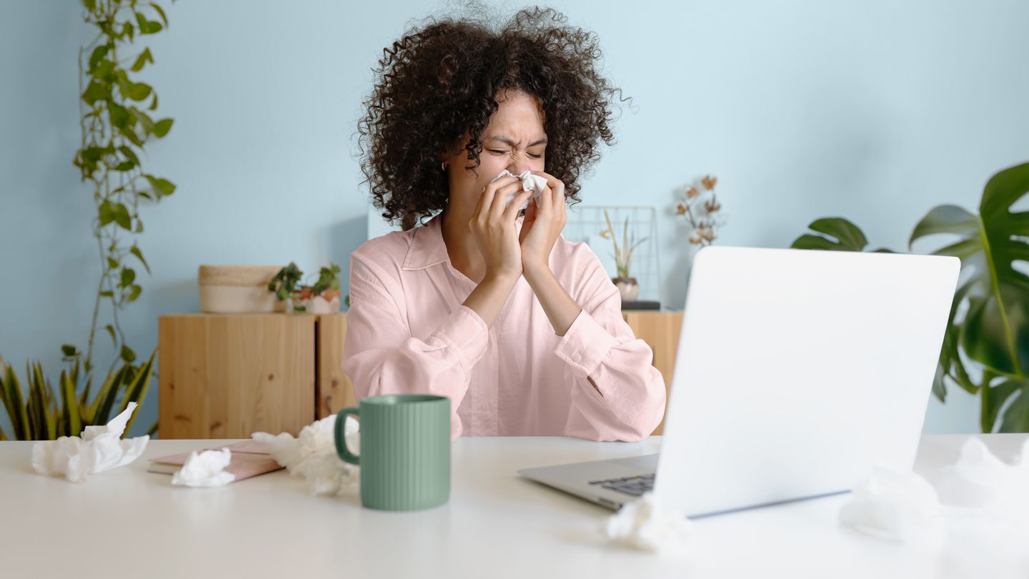young woman sitting in her home office blowing her nose with used tissues all over her desk