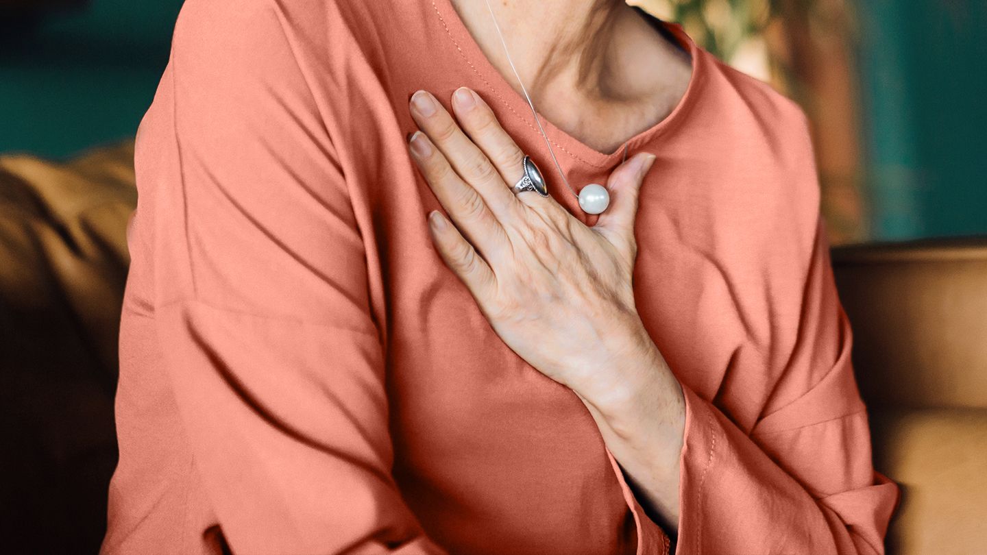 Senior Asian woman with eyes closed holding her chest in discomfort, suffering from chest pain while sitting on sofa at home.