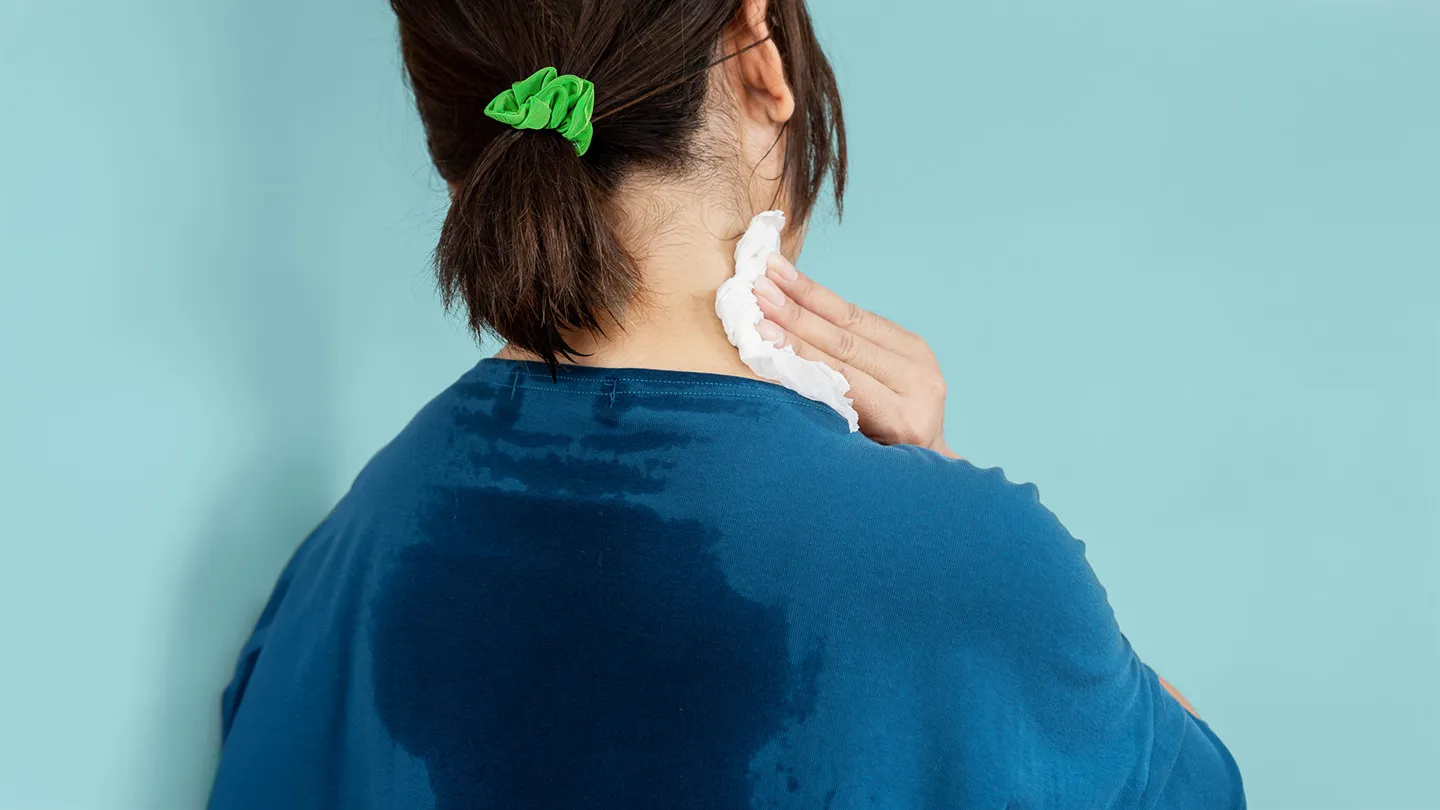 back of woman wearing green hair tie and blue tee shirt which showing excessive back sweat she is wiping sweat off neck with a tissue