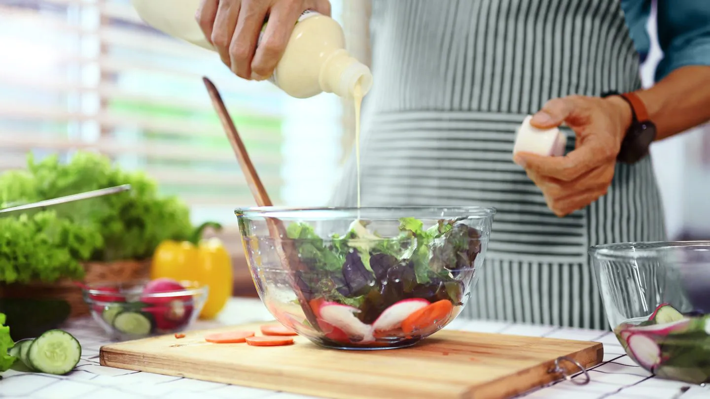 man's hands pouring salad dressing out of a jar onto bowl of salad in kitchen