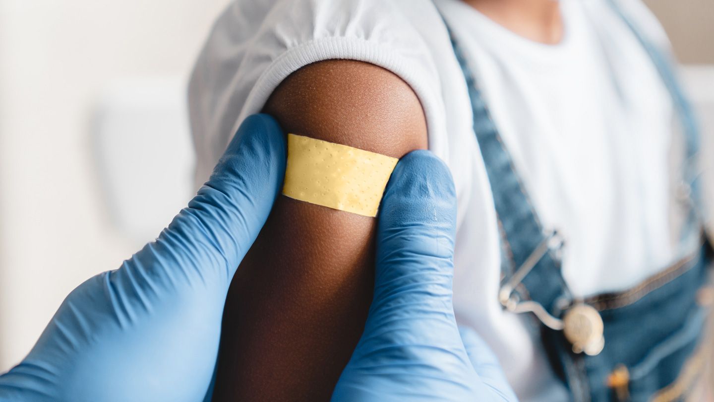 doctor's gloved hands placing yellow bandaid on young child arm after receiving the hib vaccine