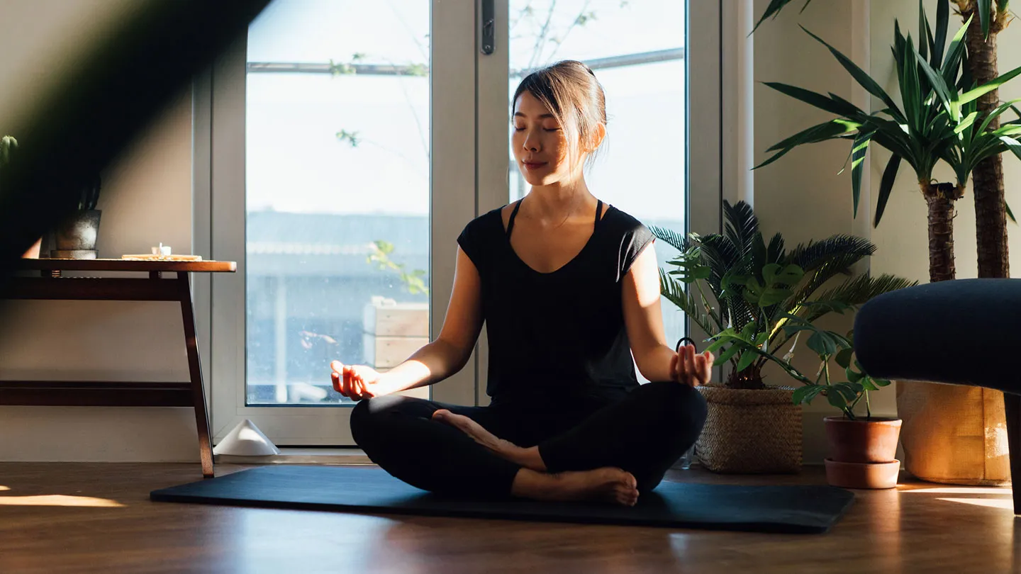 Asian woman sitting on yoga mat meditating in her livng room with eye closed surrounded by house plants