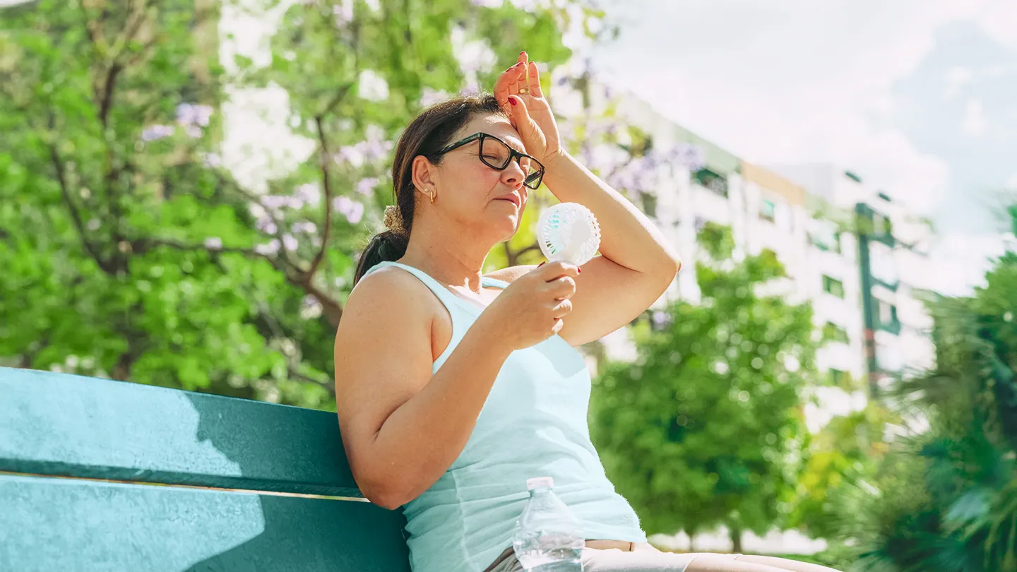 woman suffering from heat stroke and headache from extreme hot weather during heat wave holding an electric mini fan sitting outdoors on a bench with a water bottle next to her