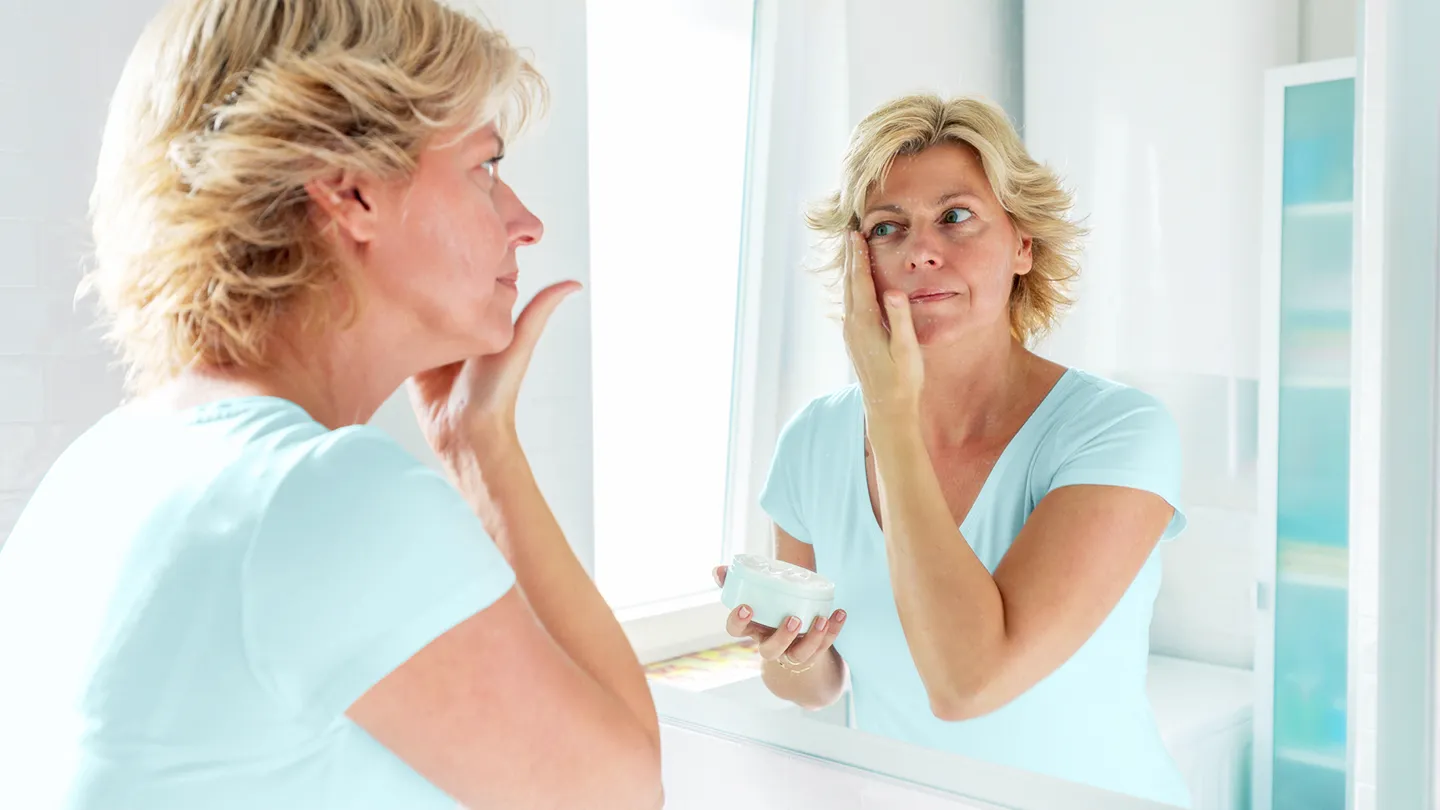 Mid adult woman in forties applying cream on her face looking in bathroom mirror