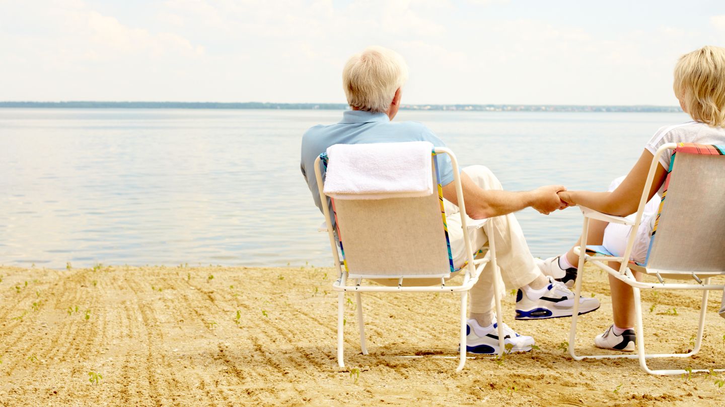 Rear view of senior couple resting in chaise lounges on lakeside and holding hands