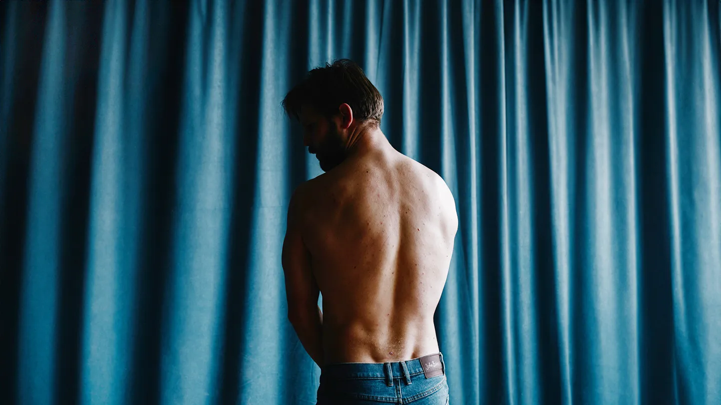 muscular man standing in shadow in front of curtain
