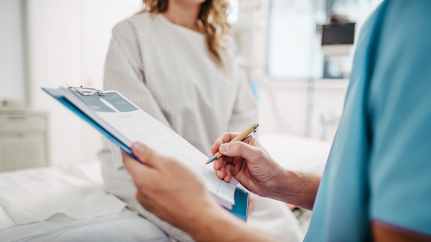 woman in patient gown sitting on table doctor in blue scrubs hands holding clipboard in front of young woman