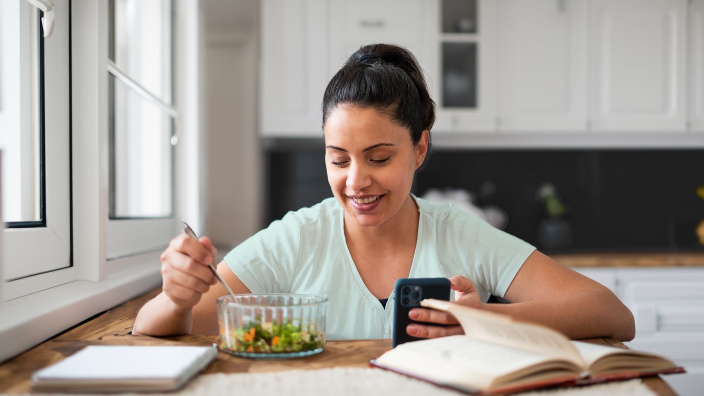 young woman looking at her phone while eating a healthy salad in her kitchen