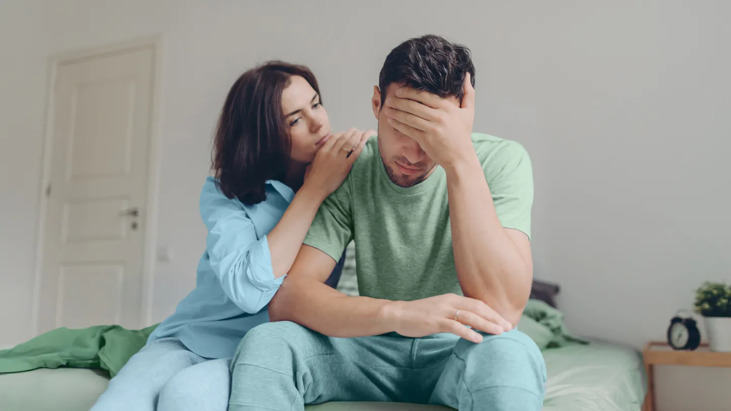 young couple sitting on their bed, woman comforting the man who is upset