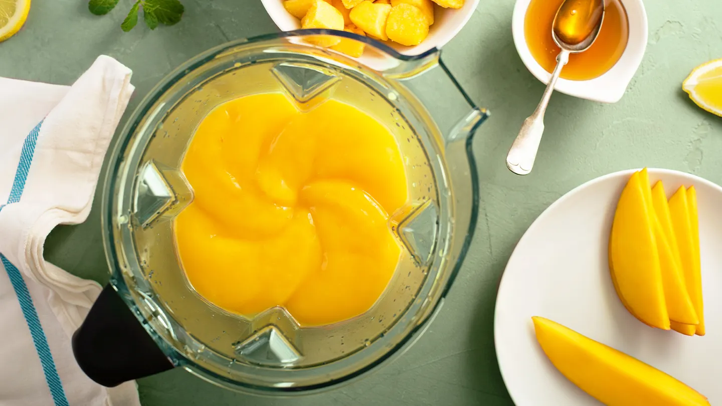 smoothie in-progress surrounded by fresh and frozen mango and cut up lemons