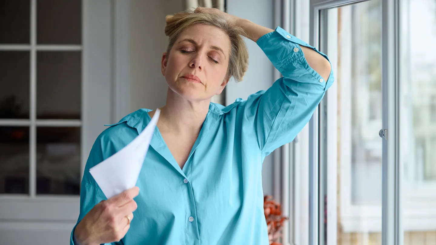 Menopausal Mature Woman Having Hot Flash At Home fanning Herself with a piece of paper