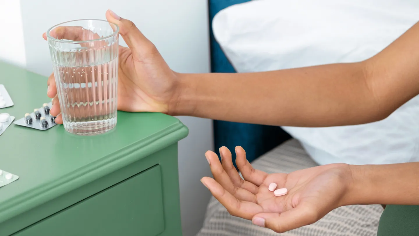 A person taking two pills with water near a bed