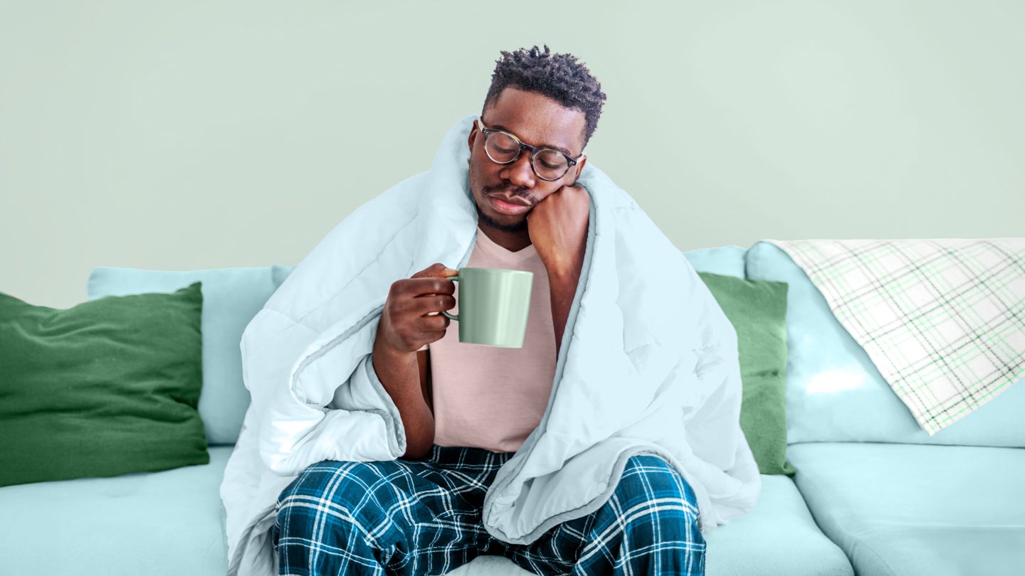 tired young Black man sitting on his couch with a comforter over his shoulders drinking coffee