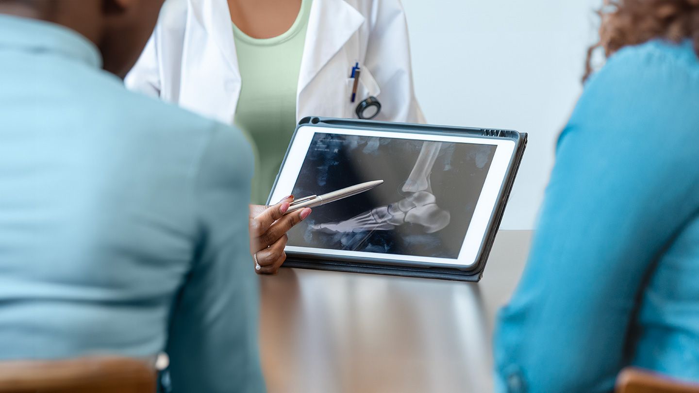 doctor pointing to the digital tablet screen of a scan for the young female patient and her mother as the doctor explains the extent the metastatic non-small cell lung cancer has spread to bones