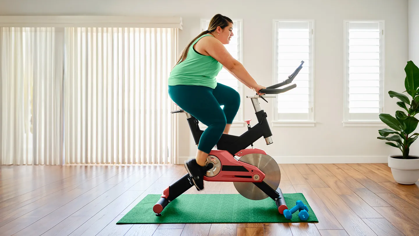 curvy woman using indoor cycling bike on mat with small weights on floor