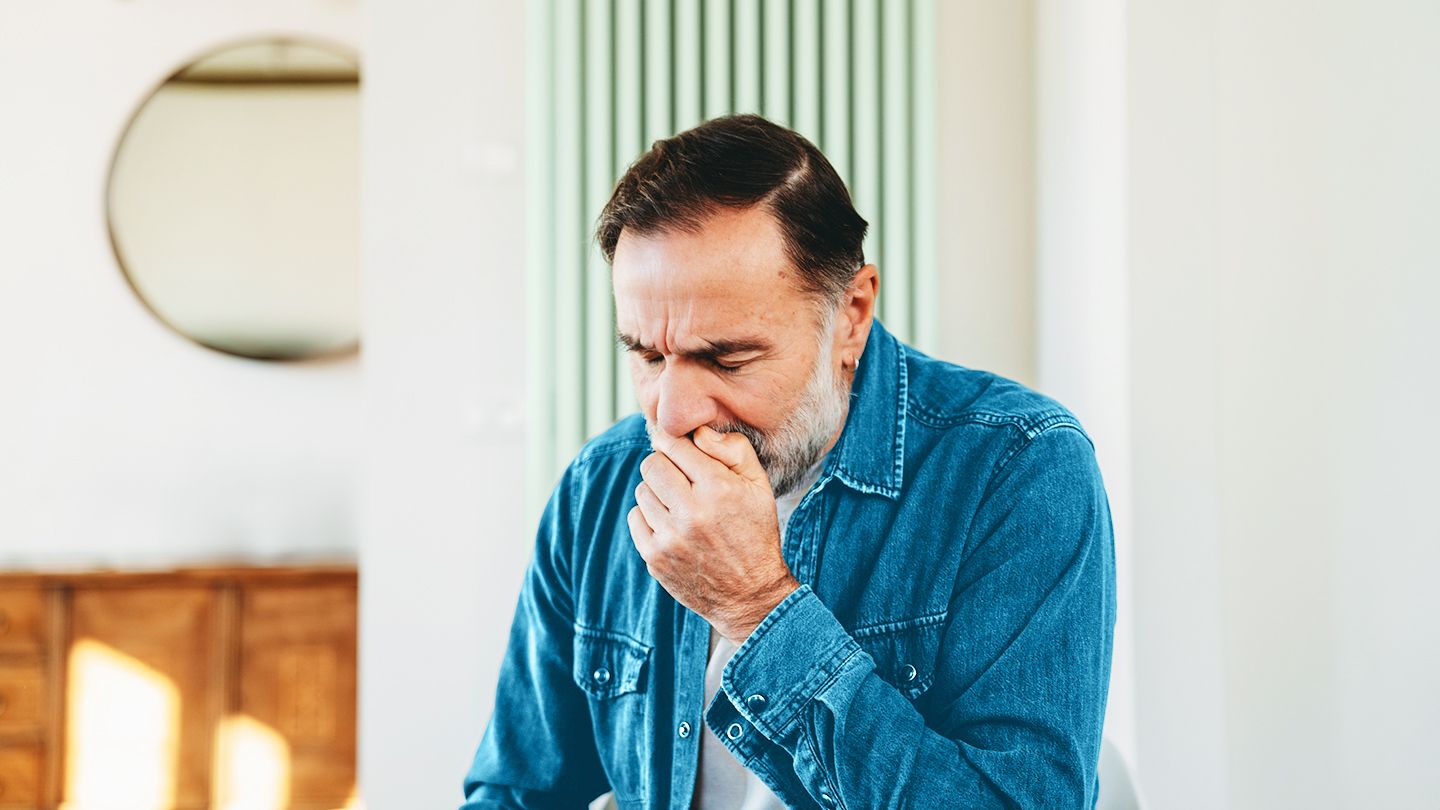 middle aged older man in blue jean jacket seated holding hand over mouth to cover dry cough