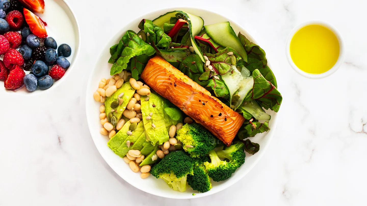 bowl of salmon leafy greens beans broccoli and cucumber bowl of berries and yogurt bowl of olive oil white countertop