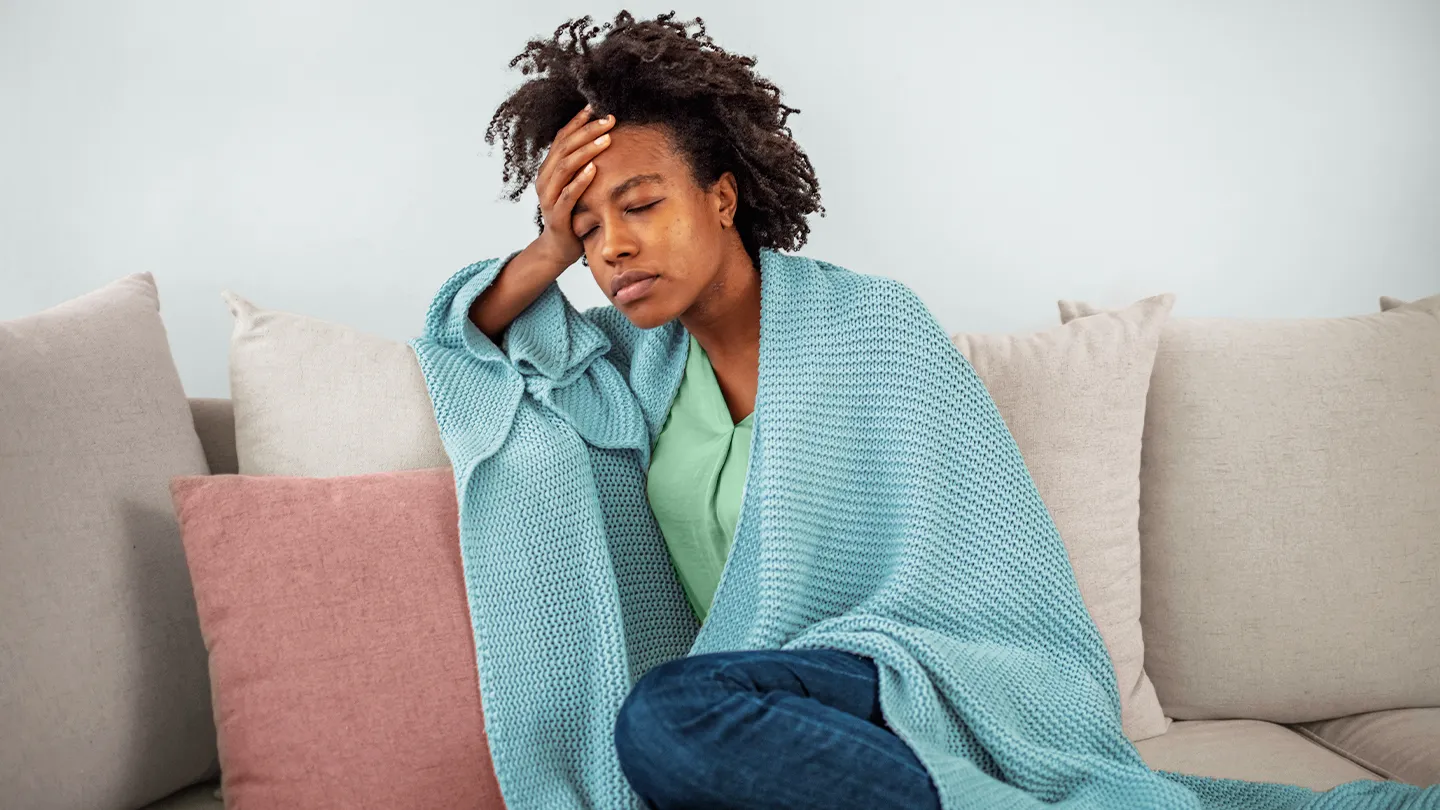 black woman sitting on couch with blanket, holding her head in pain and fatigue