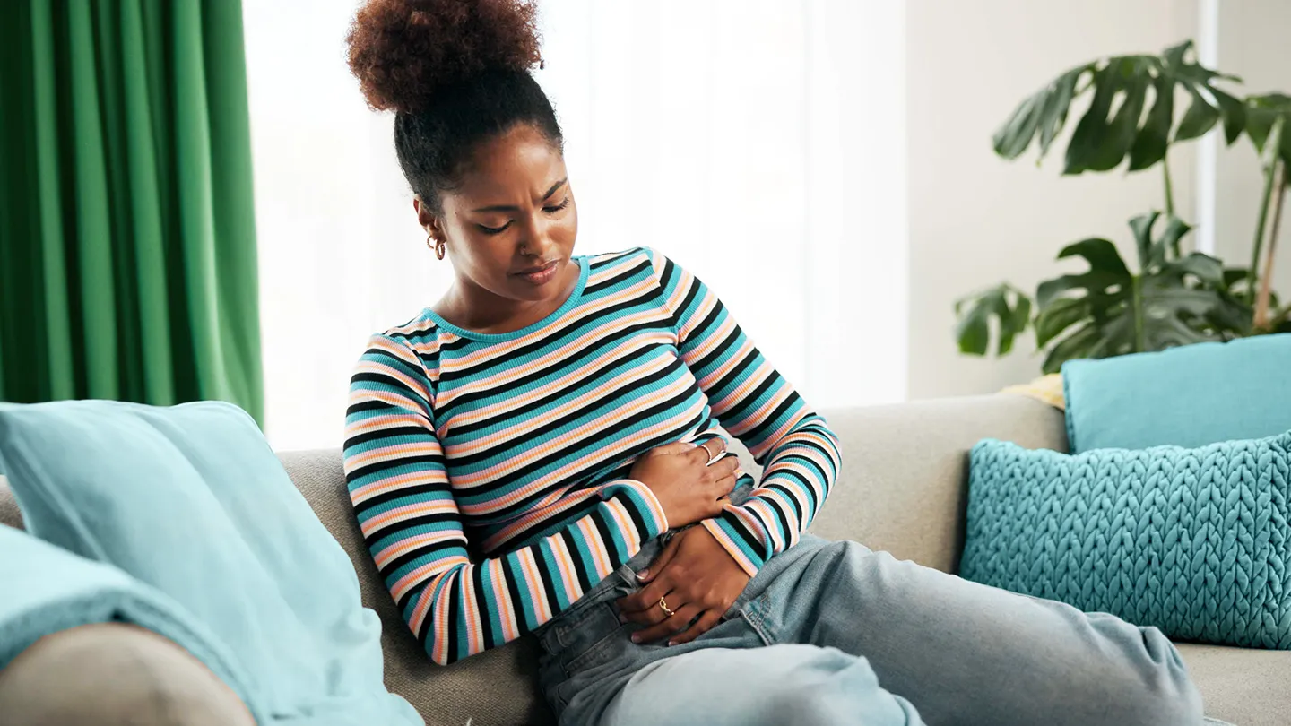 woman sitting on sofa with arms crossed over midsection with cramps and headache sensitive to the bright light coming from window behind
