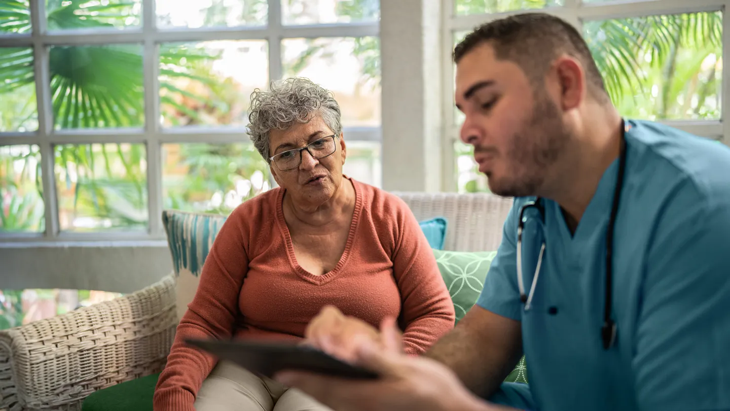 senior older adult hispanic overweight obese woman latin talking with visiting medical professional in her home on couch