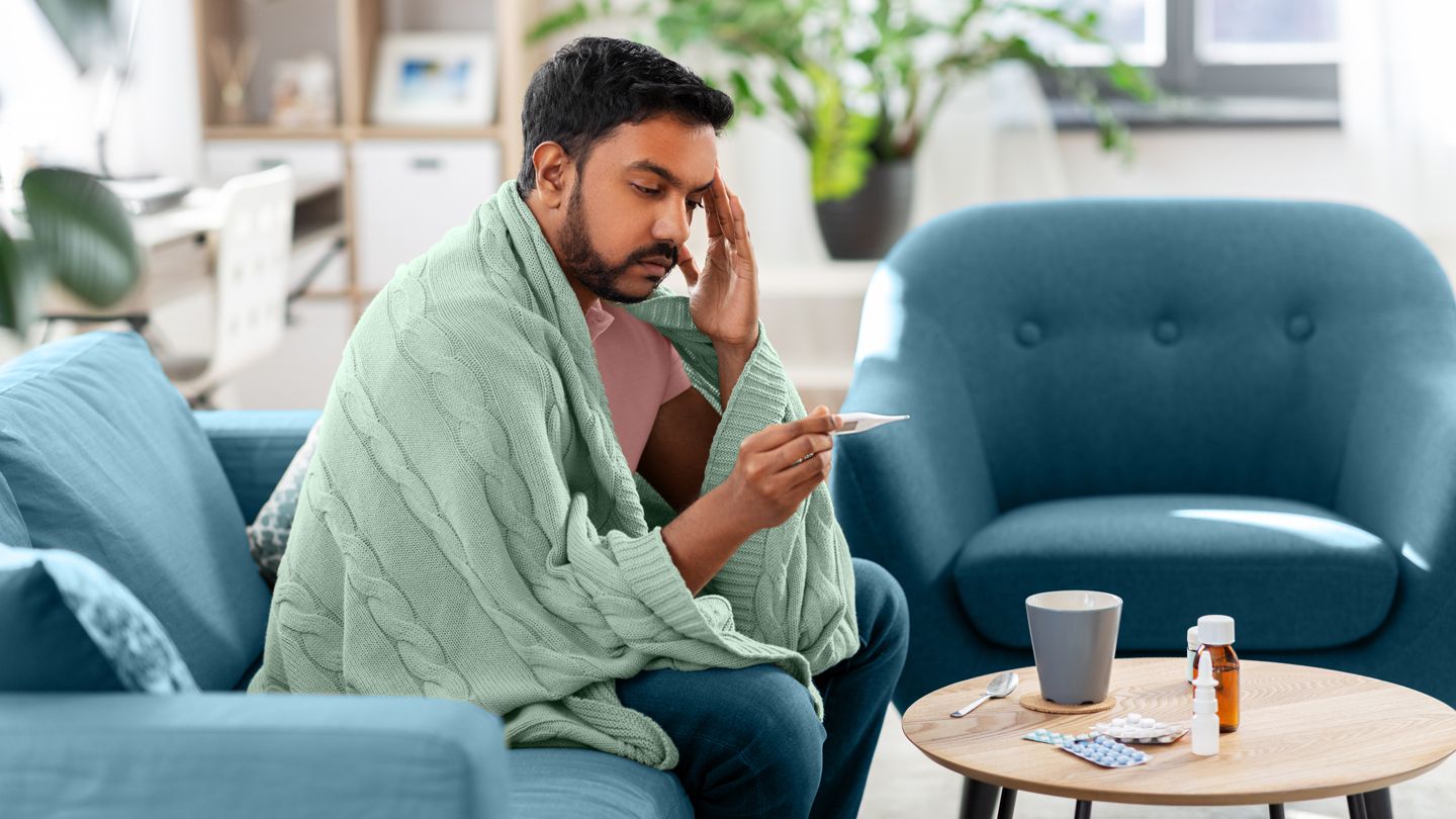 sick feverish adult man sitting on his couch with a blanket over him, taking his tempurature